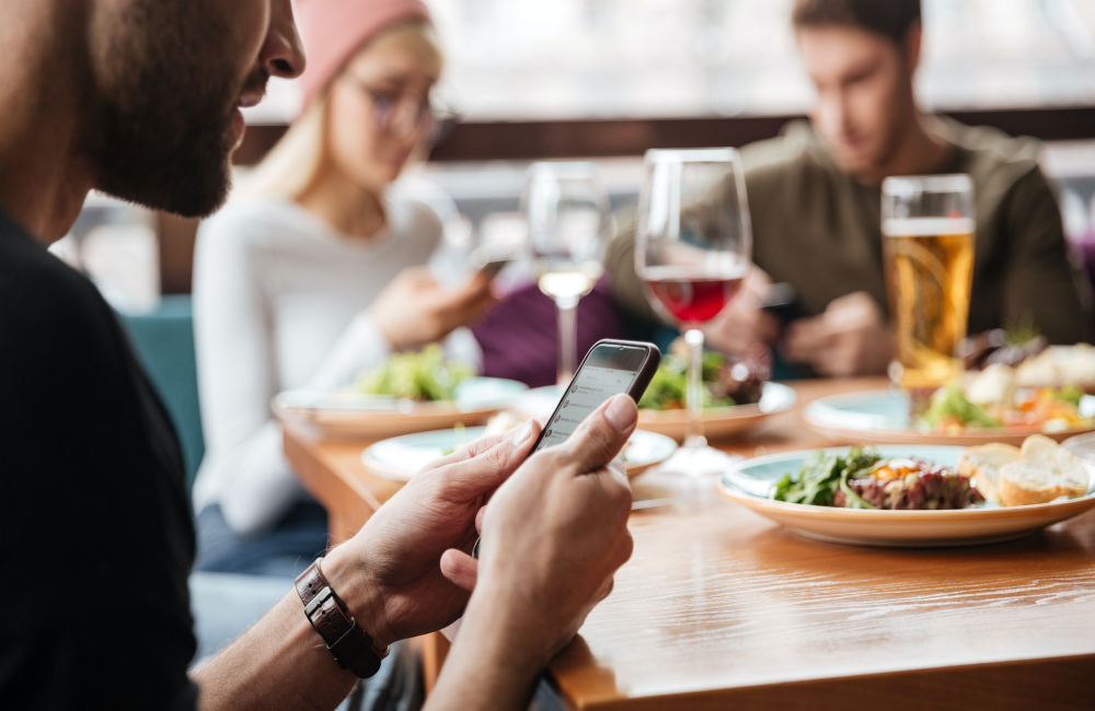 Group of men and women sitting around a table in a restaurant while shopping on their mobile phones to illustrate the concept that the word of the year in digital is "integration"