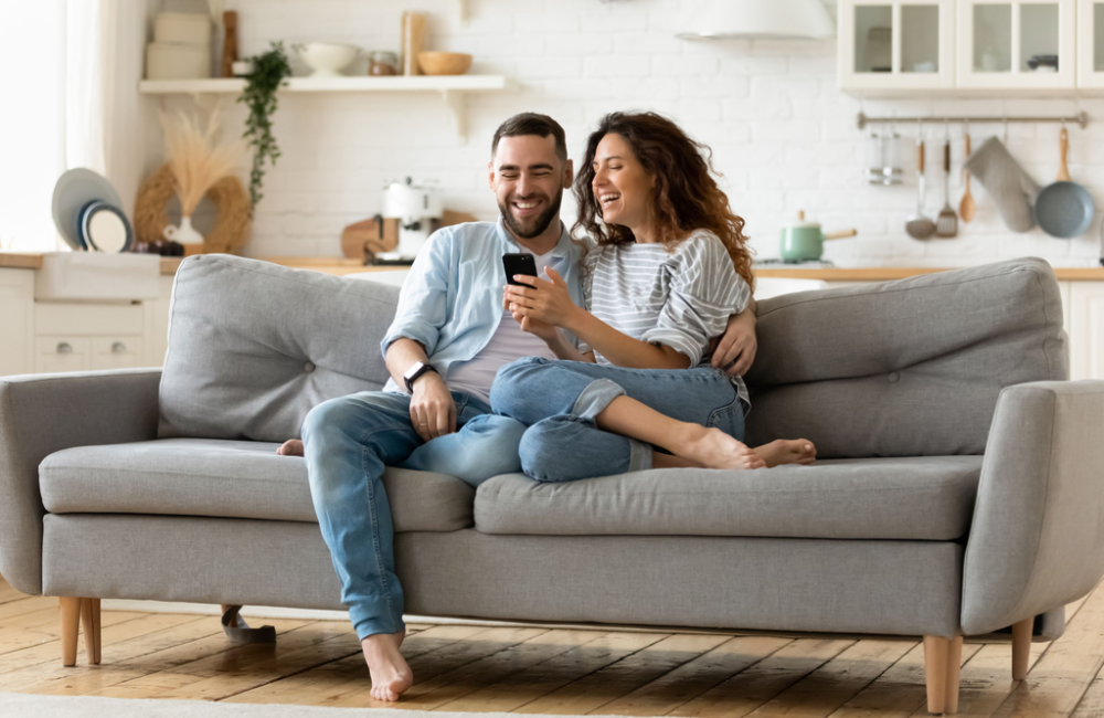 Smiling couple sitting on couch and looking at mobile phone to illustrate idea of making customers lives better